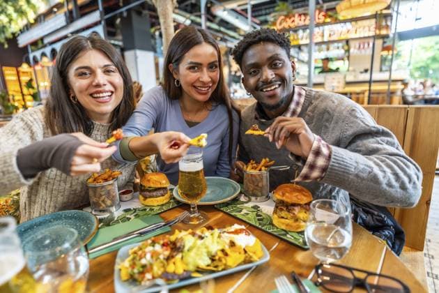Drei fröhliche, multiethnische Freunde genießen eine gemeinsame Mahlzeit mit Nachos, Burgern, Pommes und Bier in einem lebhaften Restaurant Three cheerful multi ethnic friends are enjoying a meal together, sharing nachos, burgers, fries, and beer in a vibrant restaurant setting Copyright: imageBROKER UnaixHuizi ibxuhu14216966.jpg ,model released, Symbolfoto ,property released