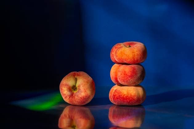 A stack of three flat peaches and one beside it sit on a reflective surface. The dark blue background enhances their vibrant colors and textures, creating a striking composition. Model Released Ellsy_ID30337_1023860_001 Copyright: xEllsyx