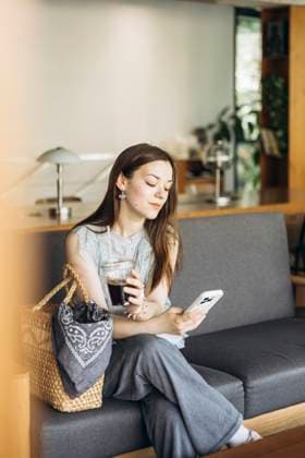 IMAGO Creative, Food, Trend, Iced Coffee A woman sits comfortably on a sofa in a cozy cafe, holding an iced coffee in one hand and a smartphone in the other, epitomizing relaxation and modern lifestyle. Model Released HannaYakimenko_ID30226_1019070_009 Copyright: xHannaxYakimenkox RECORD DATE NOT STATED