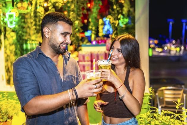 Junger Mann und Frau lächelnd und stoßen mit Cocktails in einer lebhaften Nachtmarktumgebung an, umgeben von bunten Lichtern und Grünzeug Young man and woman smiling and toasting with cocktails in a vibrant night market setting, surrounded by colorful lights and greenery Copyright: imageBROKER UnaixHuizi ibxuhu14448432.jpg ,model released, Symbolfoto ,property released