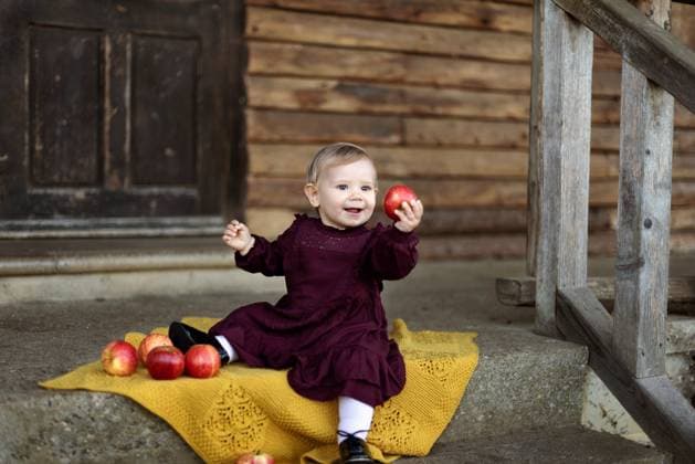 Smiling child sitting and holding an apple in front of a wooden cabin. Corby, England, United Kingdom R_JNDO250804-1797188-01 ,model released, Symbolfoto