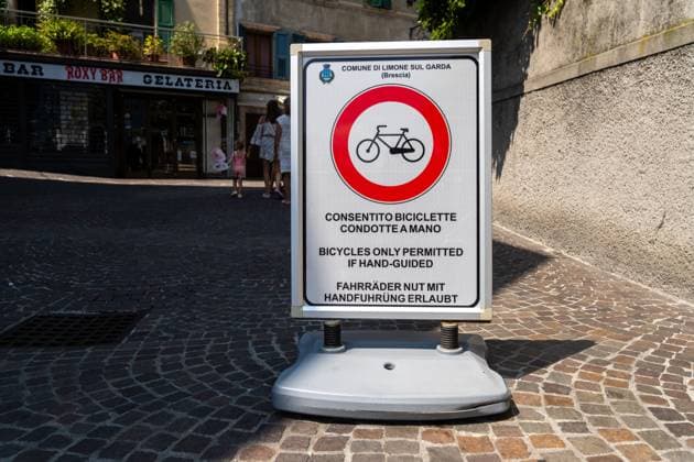 Limone sul Garda, Lombardy, Italy - August 7, 2025: Mobile traffic sign in Limone sul Garda: red circle with bicycle, note: bicycles only allowed with hand guidance. Pedestrians pass on the cobblestone square in the old town. Rule for the pedestrian zone *** Mobiles Verkehrsschild in Limone sul Garda: roter Kreis mit Fahrrad, Hinweis: Fahrräder nur mit Handführung erlaubt. Auf dem kopfsteinigen Altstadt-Platz passieren Fußgänger. Regel für die Fußgängerzone