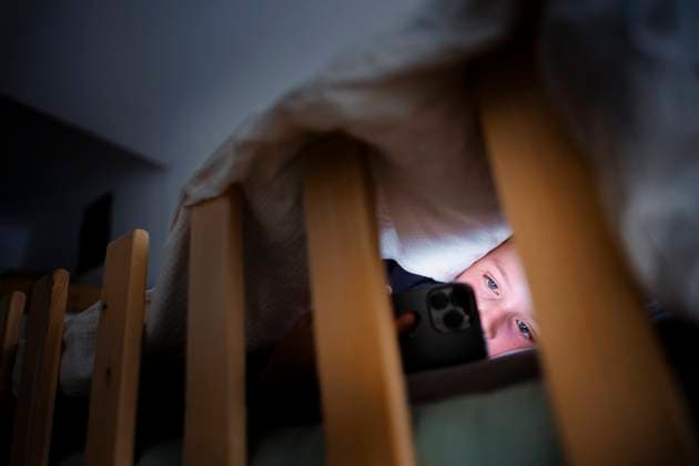 Symbolfoto: Gestellte Aufnahme zum Thema Mediennutzung bei Kindern. Ein Kind schaut im Dunkeln unter der Bettdecke im Bett auf das Display eines Smartphones. Berlin, 18.08.2025. Berlin Deutschland *** Symbolic photo Staged photo on the subject of media use by children A child looks at the display of a smartphone in the dark under the comforter in bed Berlin, 18 08 2025 Berlin Germany Copyright: xThomasxTrutschelx