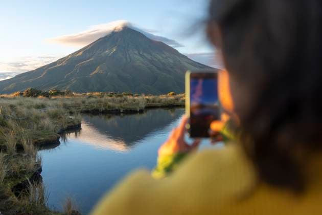 MR_ID30603_22034_MarielaWaczinsky.jpg Back view of cropped unrecognizable woman captures the stunning reflection of Mount Taranaki at sunrise in a calm lake, using her smartphone. The scene highlights nature s beauty and tranquility. Model Released JuanAizpuru_ID30603_1035172_011 Copyright: xJuanxAizpurux