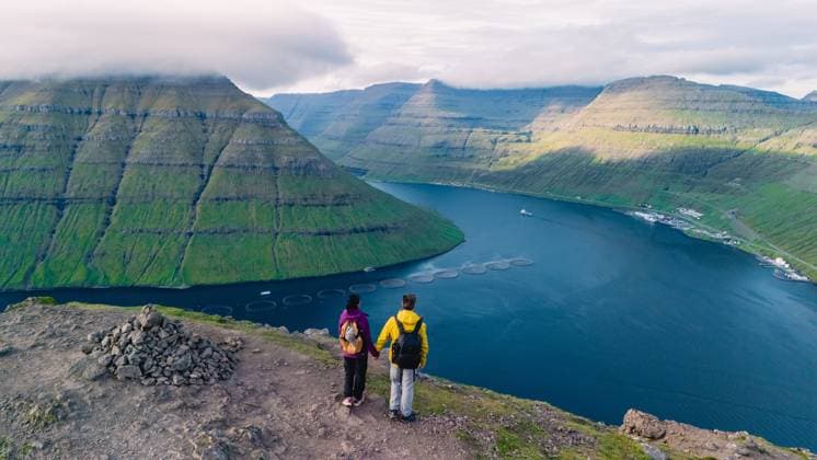 Atemberaubender Blick auf die atemberaubende Landschaft der Färöer Inseln an einem sonnigen Nachmittag Two hikers stand on a rocky vantage point Klakkur overlooking lush green valleys and tranquil waters in the Faroe Islands. The sun highlights the dramatic terrain, creating a breathtaking panorama. (License=RF) ,model released, Symbolfoto Copyright: xZoonar.com FokkexBaarssenx 23874692 ,model released, Symbolfoto ,property released