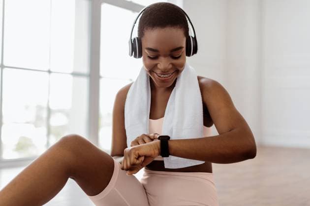 Sporty sporty lady checking fitness tracker during workout break Smiling young African American woman looking at activity fitness tracker during workout break, checking heart rate and burned calories. Happy lady wearing wireless headphones, sports bra and towel, model released, , 64239242.jpg RECORD DATE NOT STATED