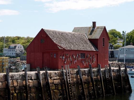 Red Wooden Building On A Stone Pier In Rockport Rockport - June, 6 2024: A red wooden building stands on a stone pier in Rockport, with a small boat in the water., 60805952.jpg RECORD DATE NOT STATED