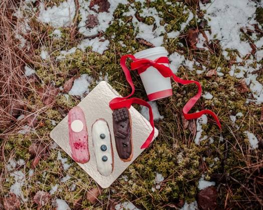 Golden Tray with Eclairs and Coffee Cup on Snowy Mossy Ground A golden tray holds three decorated eclairs beside a white coffee cup with a red ribbon, set on mossy ground partially covered with snow., 64438086.jpg RECORD DATE NOT STATED