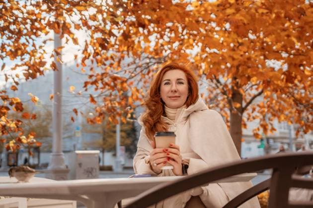 IMAGO Creative, Jahreszeit Herbst in der Stadt Woman Autumn Coffee Outdoors: Relaxed redhead enjoys fall beverage city park bench. Redhead woman smiles, holding a coffee cup, seated outdoors under vibrant autumn leaves on a park bench., model released, , 94887586.jpg RECORD DATE NOT STATED