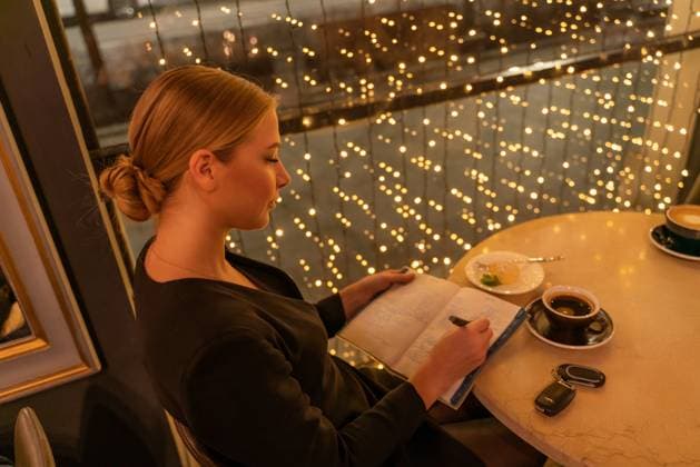 A woman is sitting at a table with a notebook and a cup of coffee. She is writing in her notebook while looking out the window. The scene suggests a moment of relaxation and contemplation. A woman is sitting at a table with a notebook and a cup of coffee. She is writing in her notebook while looking out the window. The scene suggests a moment of relaxation and contemplation, 59416748.jpg RECORD DATE NOT STATED