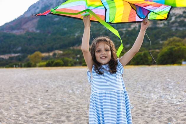 Creative Highlight Symbolbilder A young girl in a striped dress joyfully holds a colorful kite above her head on a sandy beach, with a backdrop of lush green hills under a clear blue sky. Model Released AlinaKandybka_ID31411_1068279_019 Copyright: xAlinaxKandybkax RECORD DATE NOT STATED