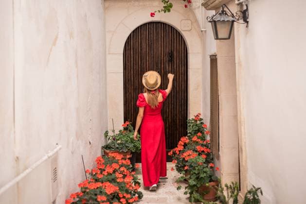 Creative Highlight Symbolbilder A woman in a vibrant red dress and a hat stands knocking on a rustic wooden door, lined with potted flowers, in an alleyway in Locorotondo, Puglia, Italy. Model Released FranciscoCarrodeguas_ID30567_1032994_046 Copyright: xFranciscoxCarrodeguasx RECORD DATE NOT STATED