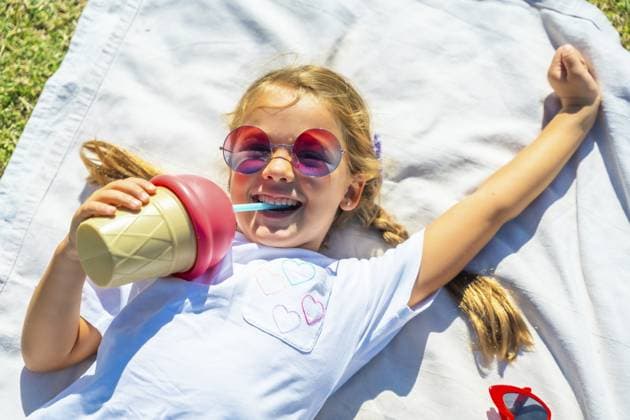 Junges Mädchen, das an einem sonnigen Tag auf einer Decke im Gras liegt und lacht, während es aus einer eiscremeförmigen Thermosflasche trinkt und bei einer Aktivität im Freien eine bunte Sonnenbrille trägt Young girl lying on a blanket in the grass on a sunny day, laughing while drinking from an ice cream shaped thermos bottle and wearing colorful sunglasses during an outdoor activity Copyright: imageBROKER UnaixHuizi ibxuhu15439935.jpg ,model released, Symbolfoto ,property released