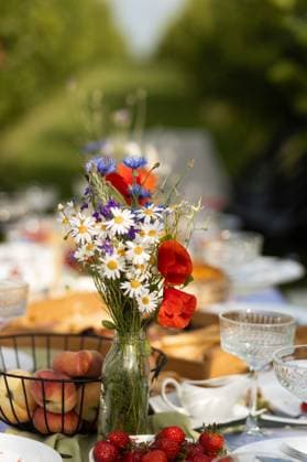 IMAGO Creative, Lifestyle, Feste im Garten #2 Floral arrangement at summer picnic brunch table A vibrant bouquet of wildflowers in a glass jar adorns an outdoor brunch table. Surrounded by peaches and strawberries, the scene exudes a warm, inviting picnic ambiance. Model Released Ptashkimenko_ID32470_1106470_001 Copyright: xPtashkimenkox RECORD DATE NOT STATED