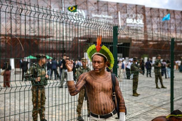 November 13, 2025, Belem, Brazil, Brazil: Amazonian indigenous man stands outside the fenced perimeter of the COP30 venue in Belem, Brazil, with military police visible inside the grounds as delegates move through the conference area. Belem Brazil - ZUMAo116 20251113_znp_o116_043 Copyright: xBiancaxOterox