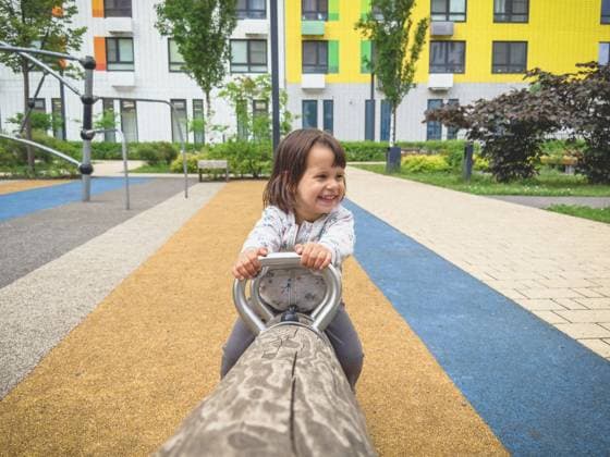 Lächelndes kleines Mädchen auf einer Wippe auf einem Spielplatz mit hellem Gummiboden und Wohnhäusern im Hintergrund, Konzept von Freude und Kindheit im Freien, Kindheitskonzept Smiling little girl on a seesaw in playground with bright rubber flooring and residential buildings in background, concept of joy and outdoor childhood, childhood concept Copyright: imageBROKER EvgeniiaxGordeeva ibxiqx15556014.jpg ,model released, Symbolfoto ,property released