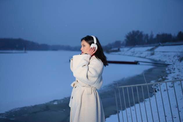 Woman in white coat with headphones by snowy river shore in winter model released, Symbolfoto, VCF01011