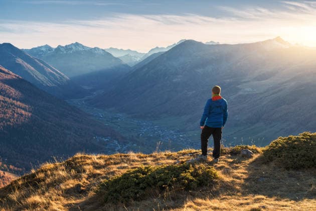 Young man standing on grassy mountain ridge with view of valley with high mountain peaks during golden hour sunset in autumn. Hiking in Italy, Livigno. Guy in Alps in fall. Trekking. Landscape Model Released DenisBilytskyi_ID32327_1102289_019 Copyright: xx