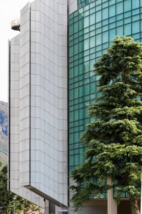 Creative Highlights World Modern building with glass facade and tree Modern building with a glass facade and geometric design, adjacent to a large green tree, showcasing urban architecture, 96460718.jpg RECORD DATE NOT STATED