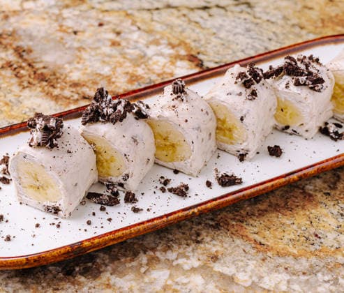 Köstliches Dessert mit gerolltem Eis und Banane, serviert auf einem Teller Six pieces of rolled ice cream with banana filling are arranged neatly on a plate. The dessert is topped with crushed cookies and placed on a textured stone countertop. (License=RF) Copyright: xZoonar.com AntonxDobreax 24631054