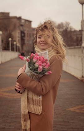 Blondine mit einem Blumenstrauß aus rosa Tulpen Blonde with a bouquet of pink tulips in the city. Girl with curls developing in the wind. She looks into the frame and smiles. Dressed in a beige classic coat. And a warm scarf, large knit. (License=RF) ,model released, Symbolfoto Copyright: xZoonar.com HannaxNikulnikovax 24130422 ,model released, Symbolfoto ,property released