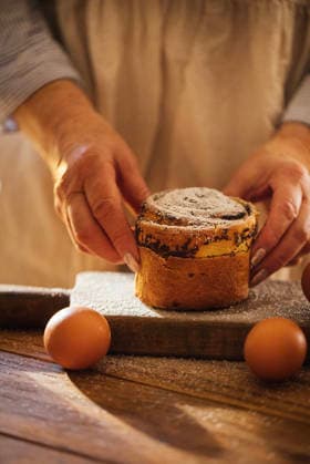 IMAGO Creative, Ostern, Bräuche, Osterbrunch Hände einer Person in einer Schürze, die einen frisch gebackenen Kuchen mit Puderzucker bestäubt auf ein Holzbrett legt, um das herum Eier verstreut liegen. Yeast dough Cruffin bun with poppy seed and succades filling. Rolled up brioche, Easter baking. Person in an apron carefully sets down a cake on a wooden board, surrounded by eggs, with a warm kitchen atmosphere in the background (License=RF) ,model released, Symbolfoto Copyright: xZoonar.com DashaxPetrenkox 24717417 ,model released, Symbolfoto ,property released