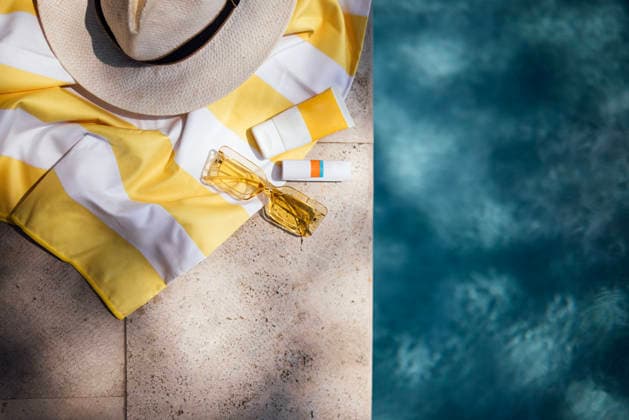 Sommer-Strandaccessoires, darunter ein Strohhut mit breiter Krempe, ein gelb gestreiftes Handtuch, eine Sonnenbrille und Sonnencreme, die auf einer Steinfläche in der Nähe eines Swimmingpools liegen. Summer beach accessories such as a straw hat, yellow striped towel, sunglasses, and sunscreen arranged on a stone surface beside a swimming pool (License=RF) Copyright: xZoonar.com DashaxPetrenkox 24726185