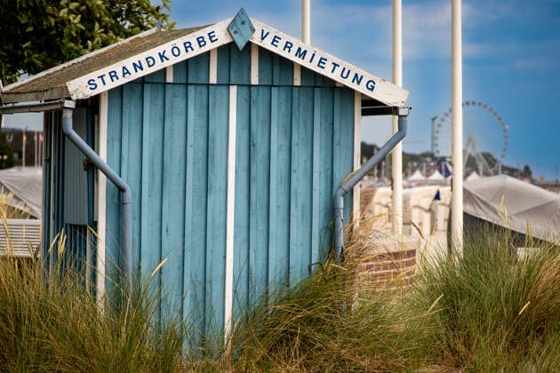 Blaue Strandkorb-Verleihhütte hinter dem Dünengras Groemitz - Schleswig-Holstein - Germany - 06-28-2025: Blue wooden hut with a beach chair rental sign stands behind dune grass, with a sandy area and a distant ferris wheel under clear sky. (License=RF) Copyright: xZoonar.com DetlefxVoigtx 24877870