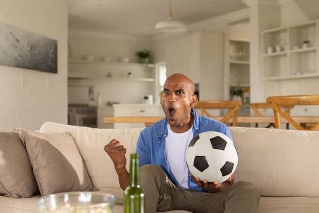 African American man sitting on sofa holding soccer ball, shouting, bottle and popcorn nearby, model released, property released, Cape Town, South Africa, 18.03.2026 06:00:00, 12882349 Copyright: xIS-260416xcreatorid:x393x 12882349