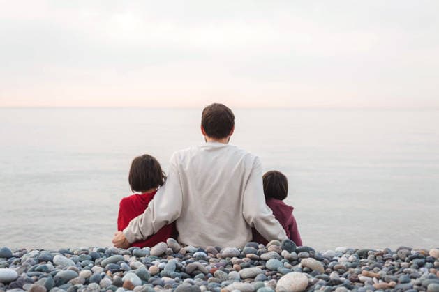 Vater sitzt mit seinen beiden Töchtern am felsigen Strand, alle sind von hinten zu sehen, er umarmt sie, während sie das ruhige Meer bei Sonnenuntergang betrachten ? ein friedlicher Familienmoment voller Liebe und Verbundenheit. Father sits on rocky beach with two daughters, all seen from behind, hugging them while watching calm sea at sunset, peaceful family moment full of love and connection (License=RF) ,model released, Symbolfoto Copyright: xZoonar.com EvgeniiaxGordeevax 25047505 ,model released, Symbolfoto ,property released