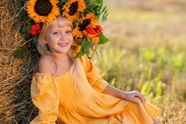 Lächelndes Kind mit Sonnenblumenkrone auf einem sonnigen Feld Smiling girl in a yellow dress sits by a haystack in a sunny field, holding a bouquet of sunflowers, radiating joy and warmth in a rural setting surrounded by hay (License=RF) ,model released, Symbolfoto Copyright: xZoonar.com MARIA_MOROZx 25056871 ,model released, Symbolfoto ,property released