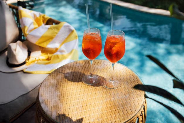 Zwei erfrischende Aperol-Spritz-Cocktails mit Orangenscheiben und Strohhalmen auf einem geflochtenen Tisch am Pool, im Hintergrund ein bunter Handtuch über einem Liegestuhl Two aperol spritz cocktails with orange slices and straws sit on a woven table next to a swimming pool, with a colorful towel on a lounge chair nearby (License=RF) Copyright: xZoonar.com DashaxPetrenkox 25113380