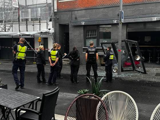 Polizisten stehen vor einem Lokal mit zerstörten Glasscheiben nach einer Explosion am Kölner Friesenwall. Im Vordergrund sind Gartenmöbel und weitere Personen sichtbar. *** Police officers stand in front of a restaurant with destroyed glass panes after an explosion on Colognes Friesenwall Garden furniture and other people are visible in the foreground