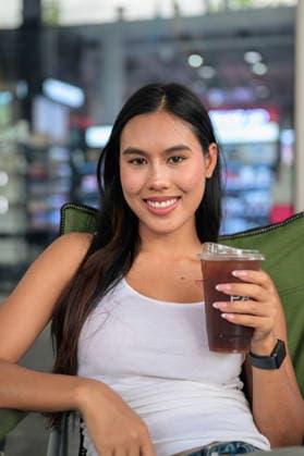 IMAGO Creative, Food, Trend, Iced Coffee Eine lächelnde Thailänderin, die einen Plastikbecher mit Eiskaffee in der Hand hält und im Freien in die Kamera blickt. Vertical medium close-up portrait of a cheerful young Isan Thai woman smiling and making direct eye contact with the camera. Seated relaxed in a green folding camping chair outdoors, she holds a clear plastic takeaway cup filled with refreshing iced coffee or cold brew against a softly blurred modern retail storefront background under bright daylight. (License=RF) ,model released, Symbolfoto Copyright: xZoonar.com TonixRantalax 25138641 ,model released, Symbolfoto ,property released
