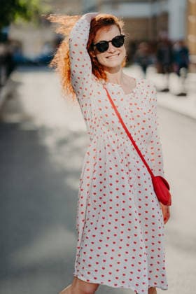 Eine rothaarige Frau trägt ein weißes Kleid mit Lippenmuster und eine Sonnenbrille und geht im Sonnenlicht durch die Straßen der Stadt Red haired woman wears white dress with lips print and sunglasses, walks on city street in sunlight (License=RF) ,model released, Symbolfoto Copyright: xZoonar.com ViorelxKurnosovx 25184582 ,model released, Symbolfoto ,property released