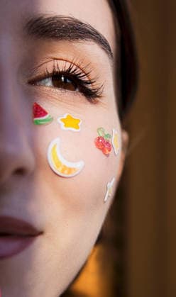 Nahaufnahme einer jungen Frau mit dekorativen Fruchtaufklebern auf der Wange, die in natürlichem Licht einen verspielten und farbenfrohen Gesichtsausdruck zeigt Young woman with fruit-themed stickers on her cheek, highlighting her facial features and playful style in soft, natural lighting (License=RF) ,model released, Symbolfoto Copyright: xZoonar.com DashaxPetrenkox 25184752 ,model released, Symbolfoto ,property released