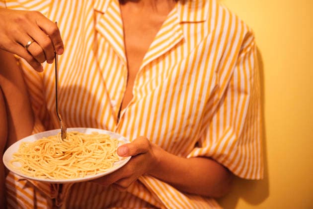 Eine Frau in einem gestreiften Pyjama sitzt an einer gelben Wand und hält einen Teller Spaghetti mit einer Gabel in der Hand; die Szene vermittelt den Eindruck eines ungezwungenen Essens zu Hause Woman in striped sleepwear enjoys a plate of spaghetti while seated against a yellow wall, creating a cozy home dining atmosphere (License=RF) ,model released, Symbolfoto Copyright: xZoonar.com DashaxPetrenkox 25184784 ,model released, Symbolfoto ,property released
