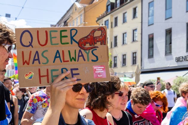 Augsburg, Bavaria, Germany June 13, 2026: Participants carrying rainbow flags and wearing colorful costumes take part in Christopher Street Day (CSD) in Augsburg. The parade through the city center promotes the rights of LGBTQ+ people as well as social diversity and tolerance. A participant holds a sign reading: QUEER WAS ALWAYS HERE *** Teilnehmerinnen und Teilnehmer mit Regenbogenflaggen und bunten Kostümen nehmen am Christopher Street Day (CSD) in Augsburg teil. Der Demonstrationszug durch die Innenstadt wirbt für die Rechte von LGBTQ+-Menschen sowie für gesellschaftliche Vielfalt und Toleranz. Teilnehmerin hält ein Schild: QUEER WAS ALWAYS HERE