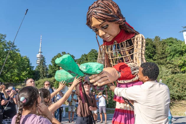 Eröffnung des Tollwood Sommerfestivals 2026, Little Amal begleitet die Festival-Eröffnung, die berühmte 3,5 Meter große Puppe, die das zehnjährige syrische Flüchtlingsmädchen Amal verkörpert, wurde weltweit zu einem Sinnbild für Hoffnung un Deutschland, München, 19. Juni 2026, Eröffnung des Tollwood Sommerfestivals 2026, Little Amal begleitet die Festival-Eröffnung, die berühmte 3,5 Meter große Puppe, die das zehnjährige syrische Flüchtlingsmädchen Amal verkörpert, wurde weltweit zu einem Sinnbild für Hoffnung und Menschlichkeit, Kinder überreichen ihr zur Begrüßung selbstgebasteltete Geschenke, im Hintergrund der Olympiaturm, das Tollwood ist multikulturelles Festival, das im Sommer im Olympiapark stattfin