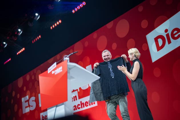Die Parteivorsitzende Ines Schwerdtner verabschiedet ihren bisherigen Co-Vorsitzenden Jan van Aken und übergibt eine Tax the Rich-Gärtnerschütze sowie ein T-Shirt mit dem Aufdruck Jan van Sprachen;Bundesparteitag Die Linke in Potsdam - Potsdamer Parteitag 2026, Potsdam Brandenburg Deutschland Metropolis-Halle *** Party Chair Ines Schwerdtner bids farewell to her former co-chair, Jan van Aken, and presents him with a Tax the Rich garden t-shirt and a T-shirt emblazoned with Jan van Sprachen. The Left Party Federal Convention in Potsdam; 2026 Potsdam Party Convention; Potsdam, Brandenburg, Germany; Metropolis Hall,detected_source_language