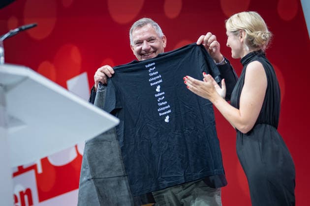 Die Parteivorsitzende Ines Schwerdtner verabschiedet ihren bisherigen Co-Vorsitzenden Jan van Aken und übergibt eine Tax the Rich-Gärtnerschütze sowie ein T-Shirt mit dem Aufdruck Jan van Sprachen;Bundesparteitag Die Linke in Potsdam - Potsdamer Parteitag 2026, Potsdam Brandenburg Deutschland Metropolis-Halle *** Party Chair Ines Schwerdtner bids farewell to her former co-chair, Jan van Aken, and presents him with a Tax the Rich garden t-shirt and a T-shirt emblazoned with Jan van Sprachen. The Left Party Federal Convention in Potsdam; 2026 Potsdam Party Convention; Potsdam, Brandenburg, Germany; Metropolis Hall,detected_source_language