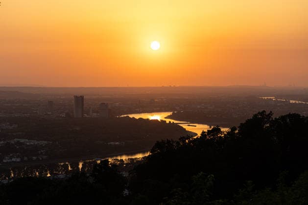 Sonnenuntergang in der Bonn Blick vom Petersberg im Siebengebirge auf Bonn bei Sonnenuntergang: Post Tower und die Hochhäuser des UN-Campus ragen aus dem goldenen Hitze-Dunst. Der Rhein mit einem Frachtschiff spiegelt das warme Abendlicht. Sommerhitze mit bis zu 35 Grad. Königswinter Petersberg Nordrhein-Westfalen Deutschland *** Sunset in Bonn: View of Bonn from Petersberg in the Siebengebirge at sunset; the Post Tower and the skyscrapers of the UN Campus rise above the golden haze of the heat; the Rhine, with a cargo ship, reflects the warm evening light; summer heat reaching up to 35 degrees; Königswinter, Petersberg, North Rhine-Westphalia Germany Copyright: xMarcxJohnx