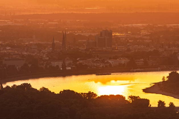 Sonnenuntergang in der Bonn Blick vom Petersberg im Siebengebirge auf Bonn bei Sonnenuntergang: Stadthaus und Bonner Münster ragen aus dem goldenen Hitze-Dunst. Der Rhein mit einem Frachtschiff spiegelt das warme Abendlicht. Sommerhitze mit bis zu 35 Grad. Königswinter Petersberg Nordrhein-Westfalen Deutschland *** Sunset in Bonn: View of Bonn from Petersberg in the Siebengebirge at sunset; the City Hall and Bonn Cathedral rise above the golden haze of the heat; the Rhine, with a cargo ship, reflects the warm evening light; summer heat reaching up to 35 degrees; Königswinter, Petersberg, North Rhine-Westphalia, Germany Copyright: xMarcxJohnx