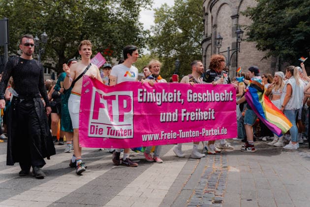 05.07.26, Cologne Pride 2026, parade and demonstration for diversity and equality Members of the Freie Tunten Partei march behind a large pink banner reading Einigkeit, Geschlecht und Freiheit (Unity, gender and freedom) during Cologne Pride 2026. The group walks past the church area with rainbow flags and dressed-up participants around them. Cologne Cologne NRW Germany