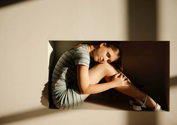 Eine junge Frau in einem gestreiften Kleid hat sich in einer rechteckigen Nische zusammengerollt; sanftes Sonnenlicht wirft Schatten an die Wand und unterstreicht die minimalistische und künstlerische Innenausstattung. Young woman with long hair sits curled up in a rectangular niche, soft sunlight creates shadows on the wall, emphasizing a minimalist aesthetic (License=RF) ,model released, Symbolfoto Copyright: xZoonar.com DashaxPetrenkox 25282711 ,model released, Symbolfoto ,property released