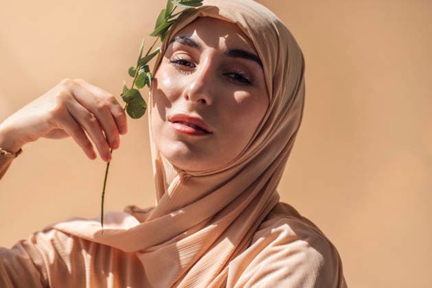 Eine junge Frau mit einem hellen Hijab hält sanft ein grünes Blatt in der Hand und strahlt vor einem sanften beigen Hintergrund bei warmer Beleuchtung natürliche Schönheit und Eleganz aus. Woman in a light hijab holds a green leaf, exuding grace and confidence, with a soft beige background that enhances her natural beauty and serene ambiance (License=RF) ,model released, Symbolfoto Copyright: xZoonar.com DashaxPetrenkox 25305536 ,model released, Symbolfoto ,property released