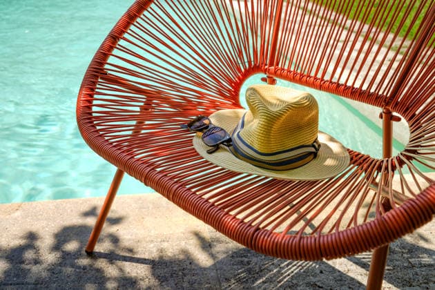 Sonnige Szene am Poolrand. Klassische Sommer-Accessoires wie ein Strohhut und eine dunkle Sonnenbrille mit markantem Rahmen auf einer Sitzbank im Acapulco-Stil *** sunny Scene on Pool edge Classic Summer Accessories like a Straw hat and a dark Sunglasses with striking Frame at a Bench at Acapulco Style 1133303198