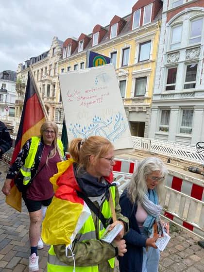 Flensburg, Schleswig-Holstein Demo ProjektM1llion öEinigkeit für Deutschland™. Hier: Person trägt Transparent mit der Aufschrift Wir ertrinken in Steuern und Abgaben, darunter bunt unterlegt Begriffe Versicherung, Fahrzeugsteuer, Grundsteuer, Umsatzsteuer, Arbeitlosenversicherung, Mehrwertsteuer, Erbschafssteuer, Pflegeversicherung, Einkommensteuer, Abgeltungs-Steuer. Dahinter eine Person mit der Deutschlandfahne in der Hand. Aufnahme vom 18.07.2026, Flensburg. *** Flensburg, Schleswig-Holstein: M1llion Unity for Germany™ demonstration. Here, a person is holding a banner that reads We’re drowning in taxes and fees, with the terms insurance, vehicle tax, property tax, sales tax, unemployment insurance, Value-Added Tax, I