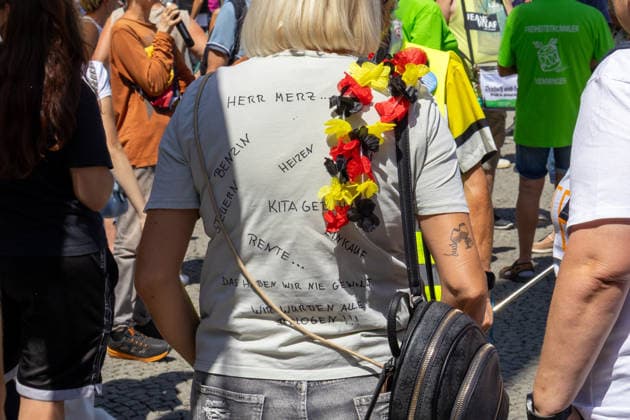 Munich, Bavaria, Germany July 25, 2026: Protesters demonstrate in downtown Munich against Chancellor Friedrich Merz (CDU) and the federal government™s policies. Among other things, the rally is directed against the government™s economic and social policies. A woman is wearing a T-shirt with the slogan: Mr. Merz... Gas, heating taxes, daycare fees, pensions, groceries. We never wanted this we were all lied to! *** Teilnehmer demonstrieren in der Münchner Innenstadt gegen Bundeskanzler Friedrich Merz (CDU) und die Politik der Bundesregierung. Die Kundgebung richtet sich unter anderem gegen die Regierungs- und Sozialpolitik. Eine Frau trägt ein T-Shirt mit Aufschrift: Herr Merz... Benzin, Heizen Steuern, Kitageld, Rente, Eink