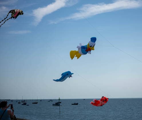 July 25, 2026, Essex, United Kingdom: Multiple novelty kites, including a cartoon-style duck, a dolphin, a tiger and a fish, fill the sky above the sea and moored boats during the Kite Festival at Southend-On-Sea, watched by a spectator on the shore below. The Kite Festival Southend s Alternative Airshow, organised by Project Southchurch, was held on the Eastern Esplanade at Thorpe Bay seafront, Southend-On-Sea, Essex. Essex United Kingdom - ZUMAs197 20260725_aaa_s197_415 Copyright: xIanxDavidsonx